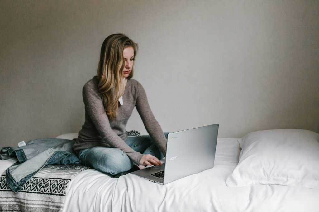 Blond woman with light skin wearing a light brown sweater and blue jeans sitting on a bed looking at her laptop.